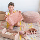Young girl holding a pink plush toy in a room with soft furnishings and toys on the floor.