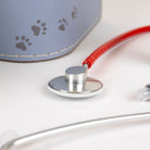 A close-up of a pet stethoscope with a silver chestpiece, red tubing, and a clear mouthpiece, shown next to a light blue pet health bag with paw print details on a white surface.