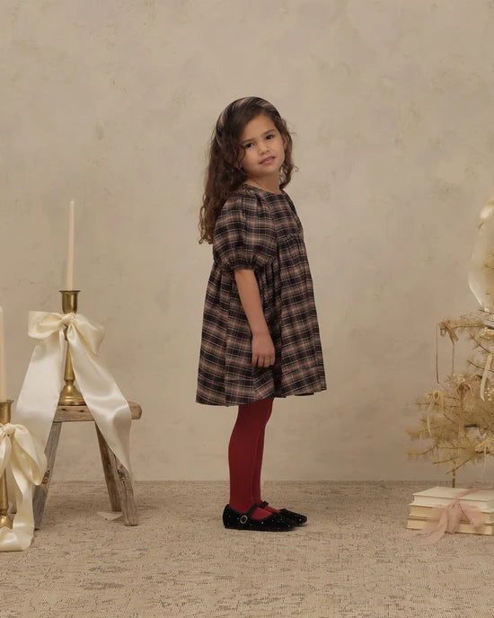 A young girl stands in a studio setting wearing a dark brown and black plaid dress, burgundy tights, and black shoes. The background features a textured beige wall and floor, decorated with a small wooden stand holding a cream ribbon and candle, and a dried floral arrangement on books to the right.