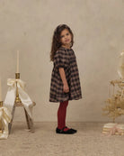 A young girl stands in a studio setting wearing a dark brown and black plaid dress, burgundy tights, and black shoes. The background features a textured beige wall and floor, decorated with a small wooden stand holding a cream ribbon and candle, and a dried floral arrangement on books to the right.