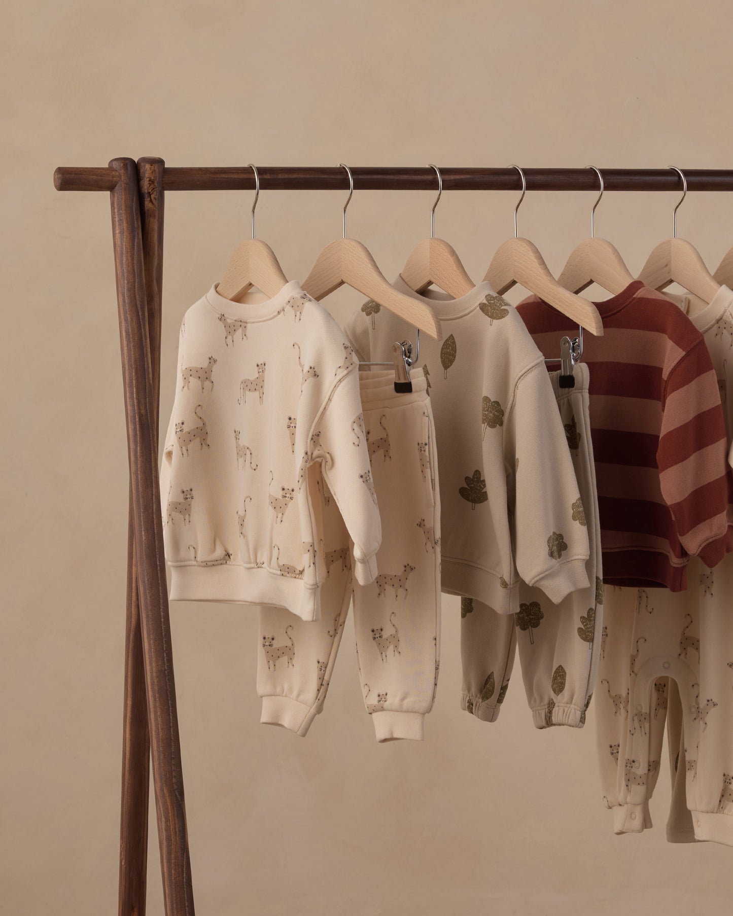 A wooden clothing rack displaying several children's two-piece sweatshirt and pants sets in neutral tones. The sets feature animal and leaf prints in beige and brown on an off-white base, with one set showing horizontal rust-red and brown stripes.