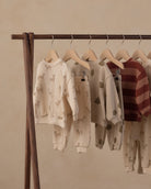 A wooden clothing rack displaying several children's two-piece sweatshirt and pants sets in neutral tones. The sets feature animal and leaf prints in beige and brown on an off-white base, with one set showing horizontal rust-red and brown stripes.