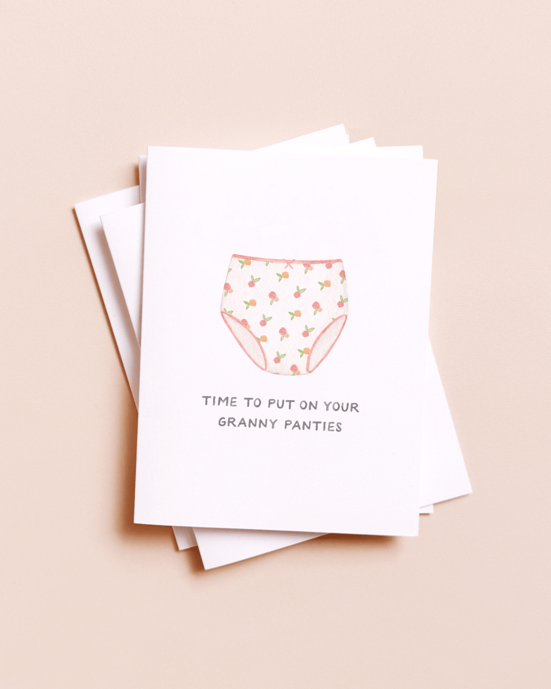 A stack of white greeting cards on a warm beige background. The top card features a watercolor-style illustration of granny panties with a pink and green floral pattern, and the text 'TIME TO PUT ON YOUR GRANNY PANTIES' printed below.