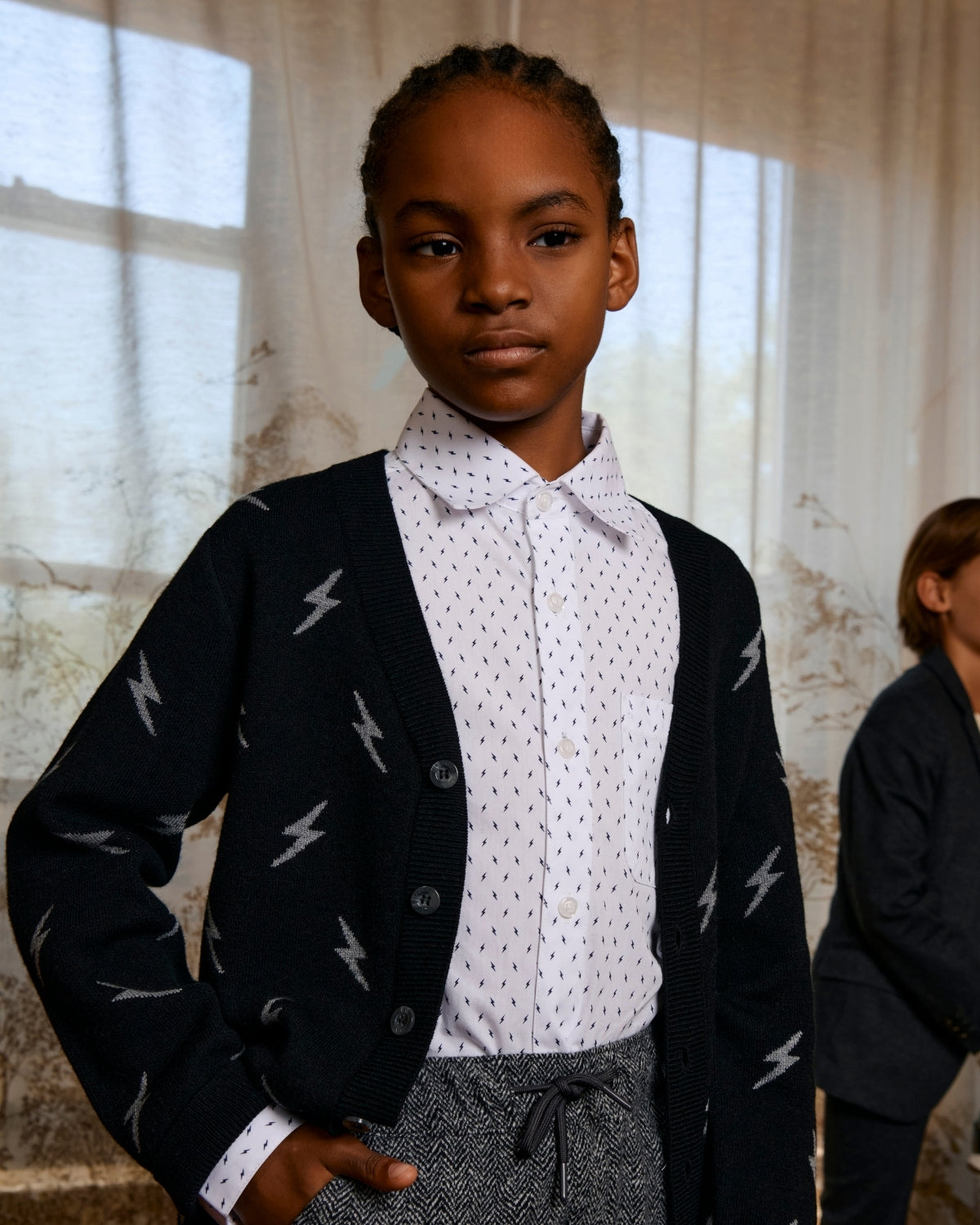 Young person wearing a black cardigan with white patterns, white shirt, and gray pants in a softly lit room.