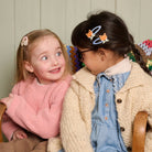 Two young girls sitting together, smiling and looking at each other, with decorative hair clips on their heads.