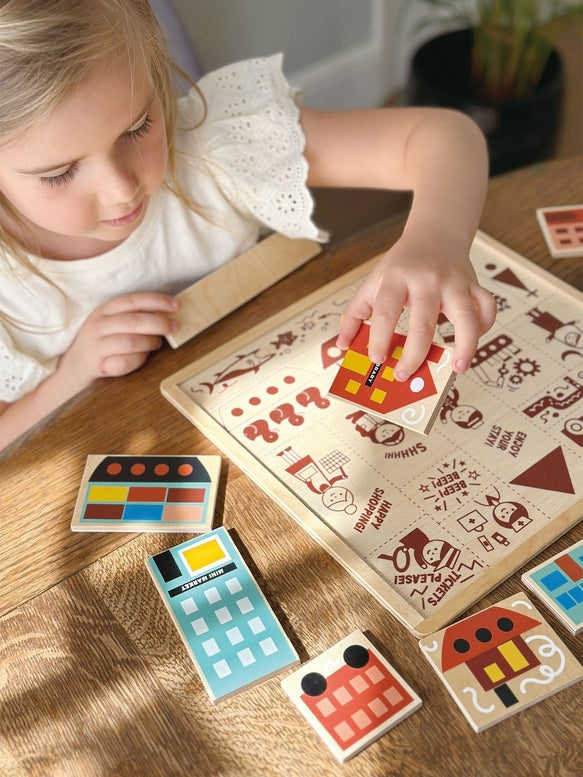 A young girl with blonde hair tied back sits at a wooden table, smiling as she plays with the Mentari Wooden Town Puzzle, holding a puzzle piece in her hand while assembling the colorful town scene.
