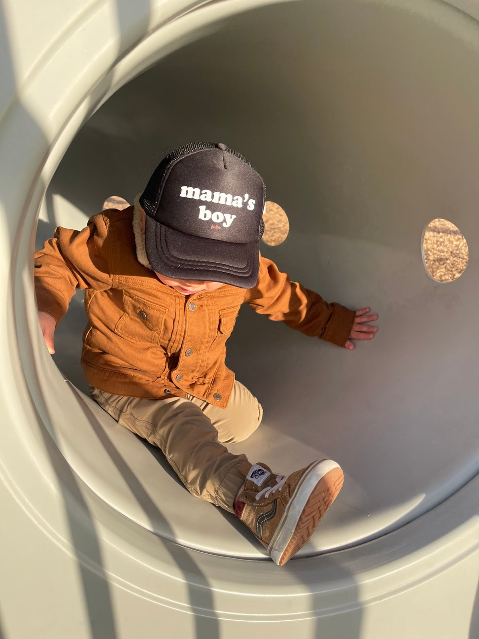Child wearing a 'mama's boy' cap sitting inside a large concrete pipe.