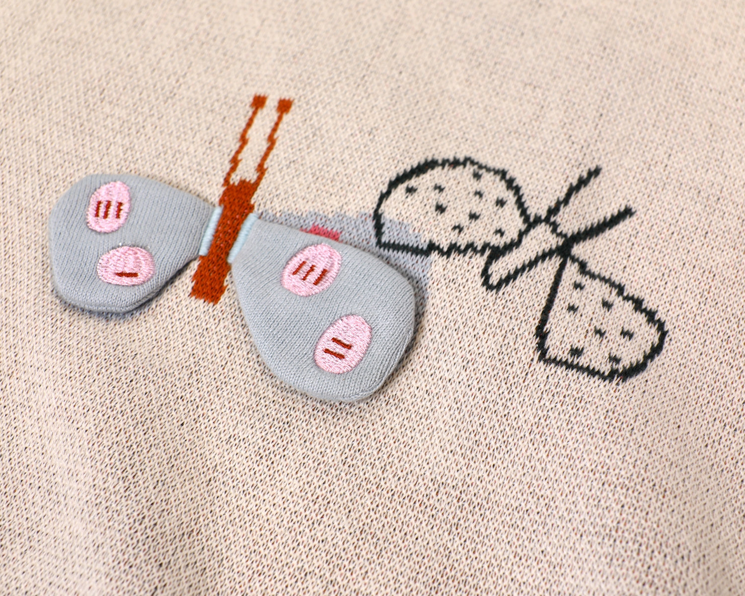 A close-up view of a light pink and white knit baby blanket, featuring a prominent 3D light blue butterfly motif with orange antennae, pink accents, and a smiling face, alongside an embroidered black-and-white butterfly outline.