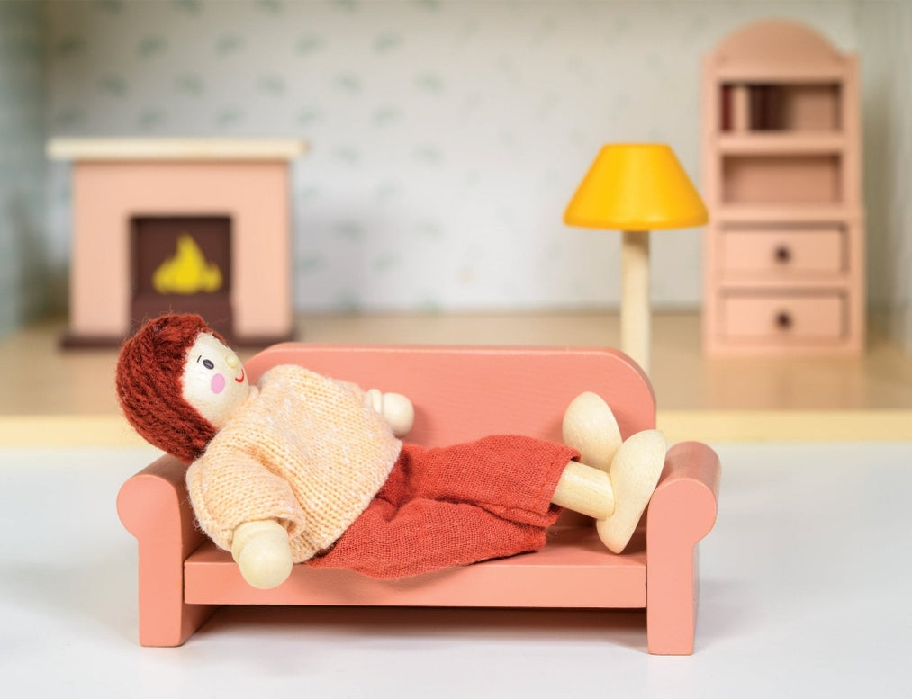 A close-up shot of a doll resting on a coral-pink toy sofa, wrapped in a knitted blanket, with a miniature wooden fireplace, a yellow floor lamp, and a bookcase in the background.