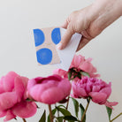 Hand holding a card with blue dots next to pink flowers on a light background