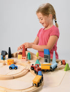 A young child with blonde hair tied back, wearing a pink short-sleeve shirt, reaches over a wooden toy train set laid out on a light-colored surface to place a red toy car onto the tracks. The setup features a figure-eight track configuration with a blue train engine and cargo cars, alongside colorful wooden town elements, including houses, trees, traffic lights, and a bridge.