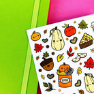 A sheet of autumn-themed stickers featuring cute cartoon illustrations of pumpkins, acorns, leaves, and pie slices on a white background, styled on top of bright green and pink paper sheets.