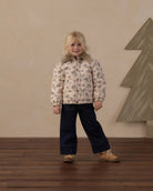 A young girl stands in a studio setting wearing a cream-colored puffer jacket with a gray floral pattern, dark navy wide-leg pants, and tan boots. The background features a textured beige wall and a large gray geometric tree decoration on the right.