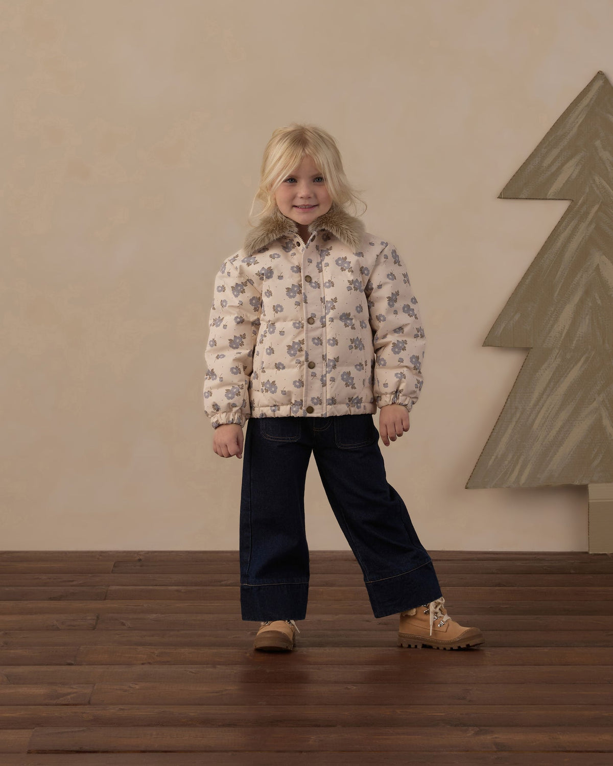 A young girl stands in a studio setting wearing a cream-colored puffer jacket with a gray floral pattern, dark navy wide-leg pants, and tan boots. The background features a textured beige wall and a large gray geometric tree decoration on the right.