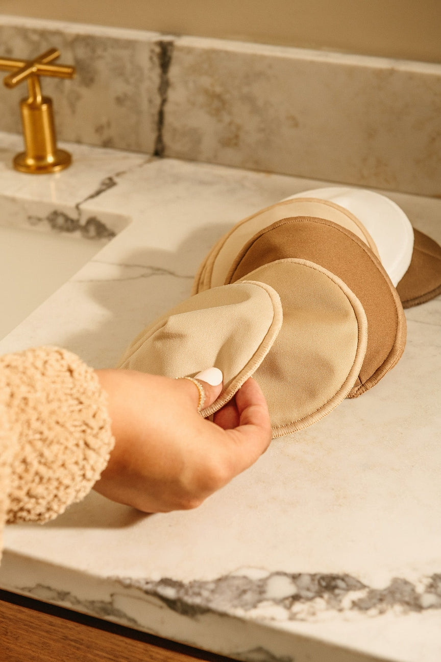 Person holding a beige bath pouf on a marble countertop with a gold faucet in the background.