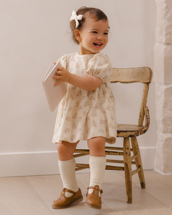 A toddler girl wearing a cream-colored short-sleeve dress with a delicate floral pattern, standing next to a rustic wooden chair and holding a book.