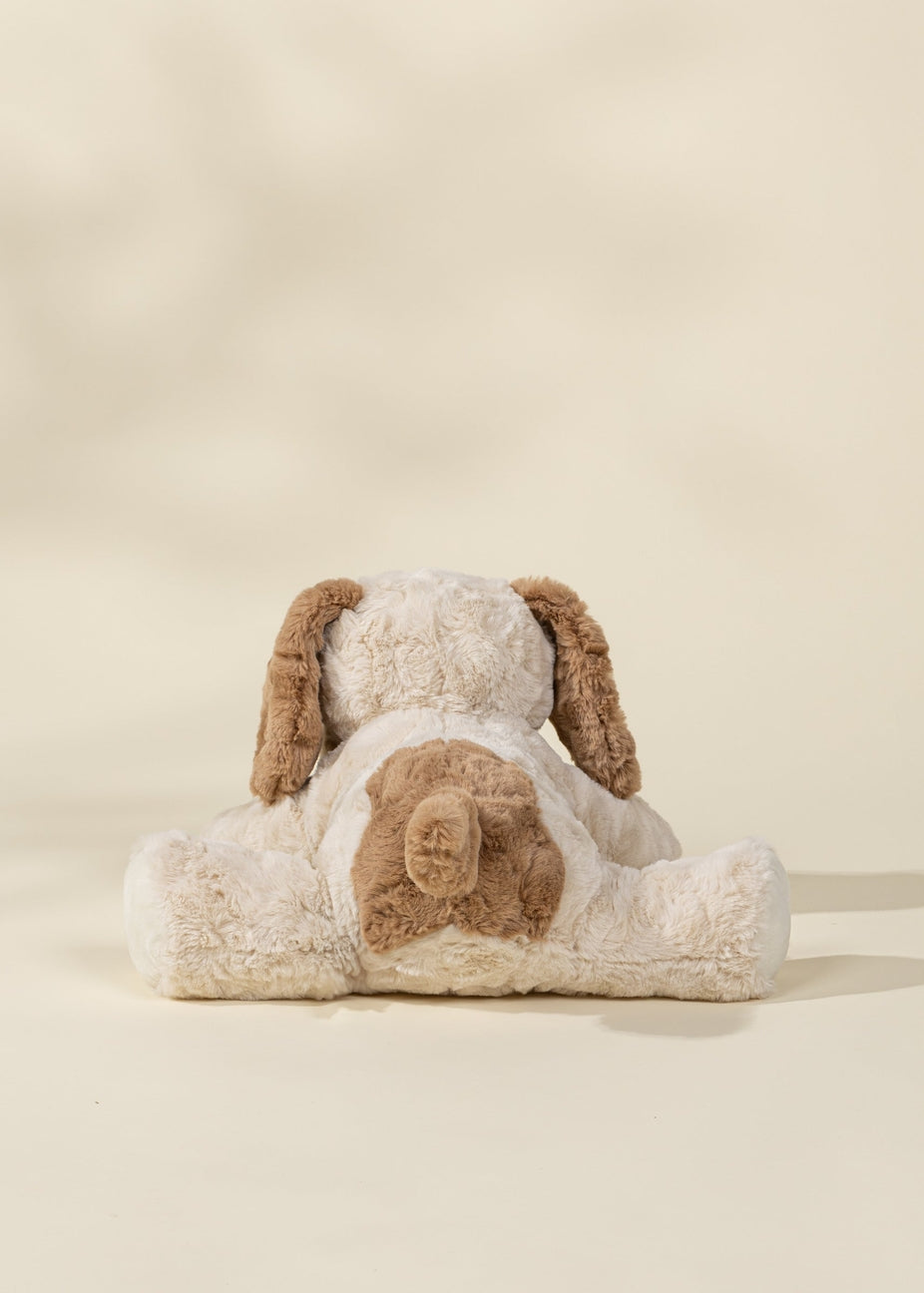 A plush stuffed dog toy shown from the back, featuring cream-colored fur with brown floppy ears and a brown patch on the back, set against a warm beige background.