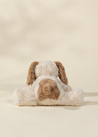 A plush stuffed dog toy shown from the back, featuring cream-colored fur with brown floppy ears and a brown patch on the back, set against a warm beige background.
