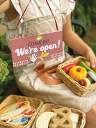 Person wearing an apron with a 'Come in! We're open!' sign, surrounded by toy food items.