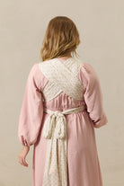 A woman shown from the back wearing a dusty pink long-sleeve dress with a cream patterned wrap belt tied around her waist, set against a plain beige studio background.
