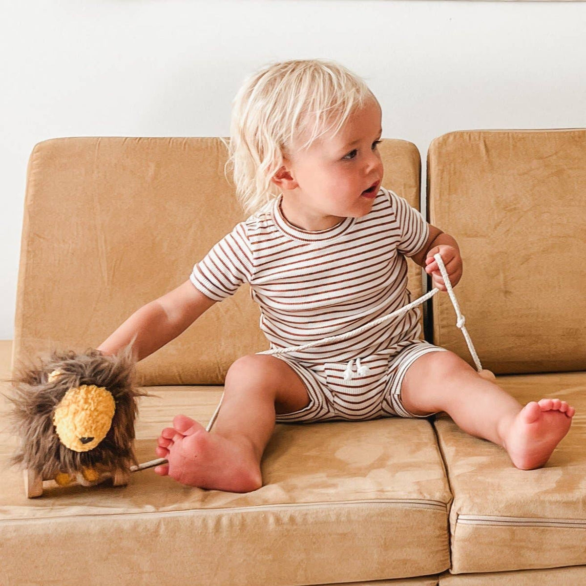 Child sitting on a beige couch holding a toy, wearing a striped outfit.