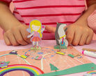 Child playing with the Floss & Rock Magnetic Scene & Doodle Set in the Rainbow Fairy theme, setting up magnetic characters like a fairy and a seal on a bicycle on a colorful magnetic board.