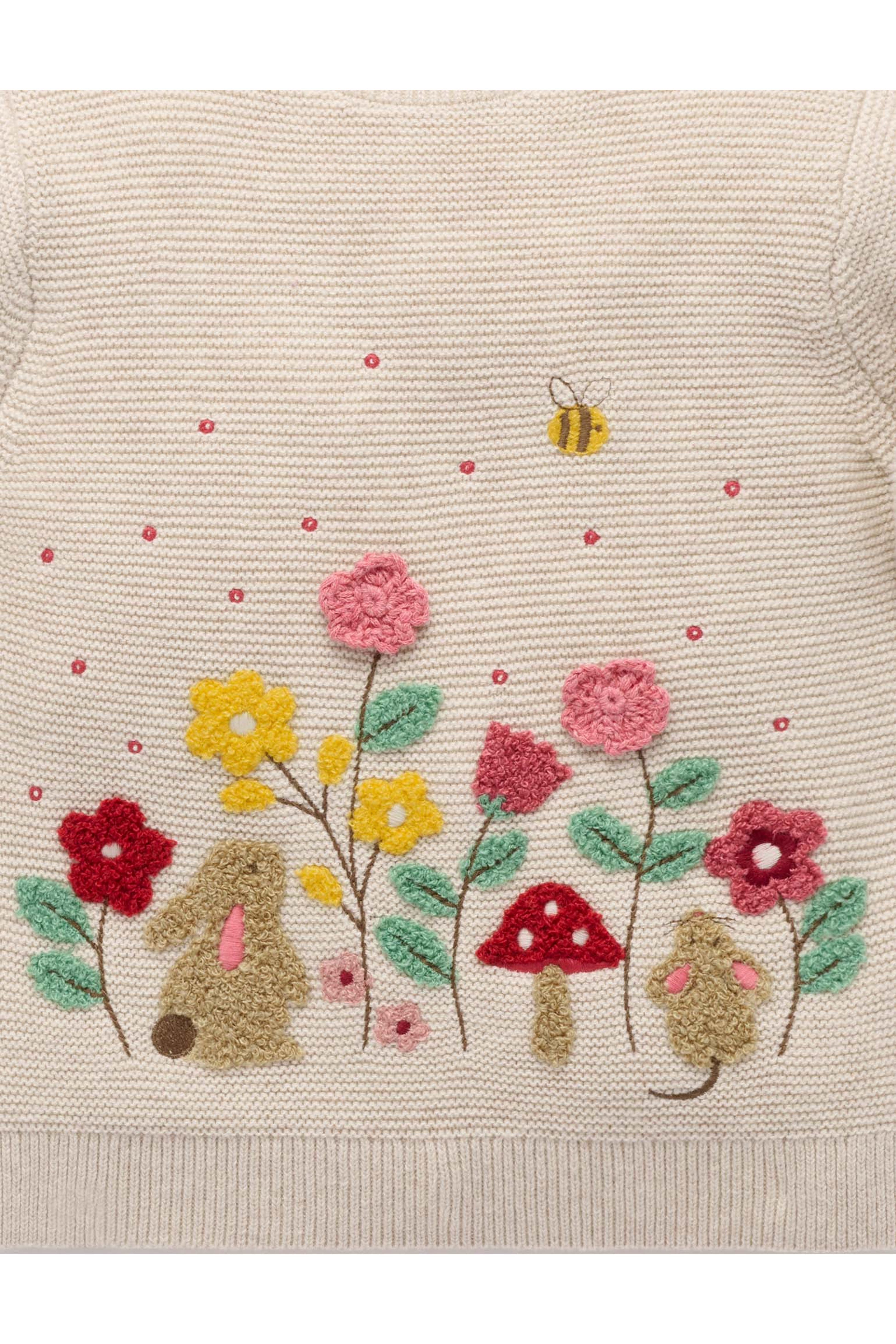 Close-up of a cream-colored knit sweater featuring colorful embroidered wildflowers, mushrooms, bunnies, a bee, and small pink dots.