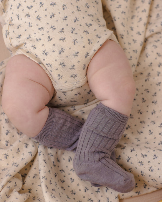 A close-up of a baby's legs wearing ribbed mauve-purple knit booties, resting on a cream-colored floral blanket.