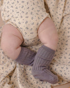 A close-up of a baby's legs wearing ribbed mauve-purple knit booties, resting on a cream-colored floral blanket.
