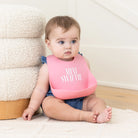 Baby wearing a pink bib with 'MINI SWIFTE' text, sitting on a bed.