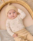 A baby lying on their back inside a woven moses basket, wearing a cream-colored long-sleeve wrap bodysuit with a small floral print, a matching cream knit hat, and tan pants.