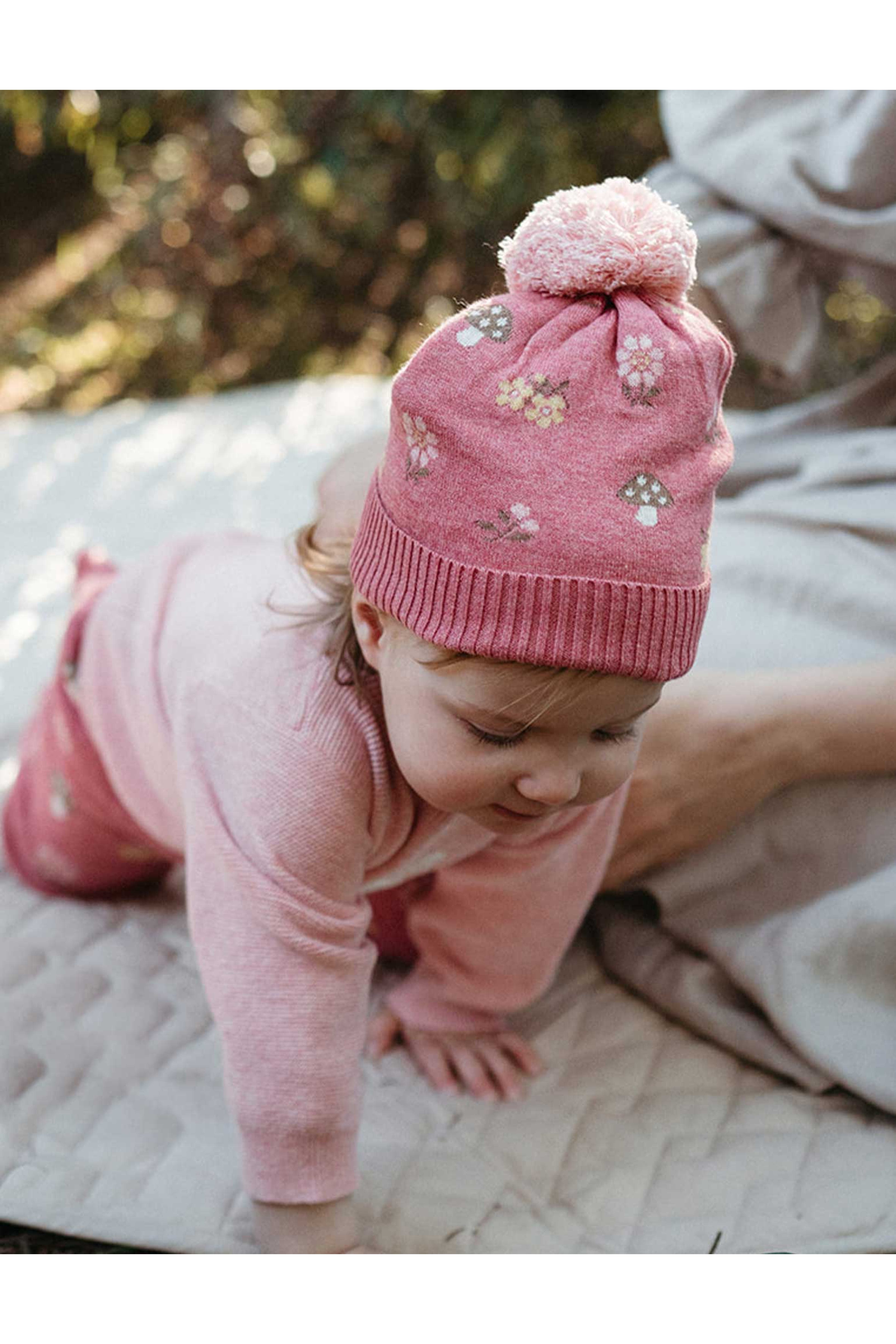 A baby wearing a pink knit hat with a pom-pom and embroidered floral pattern, crawling on a quilted blanket outdoors.