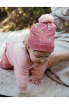 A baby wearing a pink knit hat with a pom-pom and embroidered floral pattern, crawling on a quilted blanket outdoors.