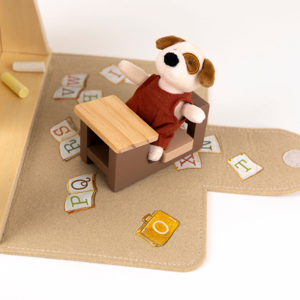 A small plush dog toy sits in a brown and tan desk chair on a beige fabric play mat, surrounded by scattered alphabet letter tiles and a few small accessories.