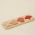 A wooden four-piece nesting shell puzzle toy with four organic-shaped wooden pieces in light pink, dusty rose, terracotta red, and bright red, set on a light natural wood rectangular board against a warm cream background.