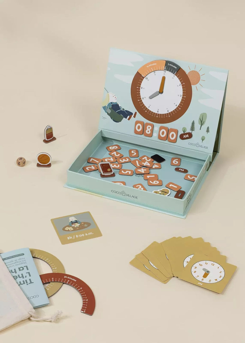 An open light blue game box labeled 'COCO VILLAGE' on a cream background, surrounded by game pieces, cards, a protractor, dice, and a small bag. The box lid shows a clock illustration and the inside contains many small game tokens.