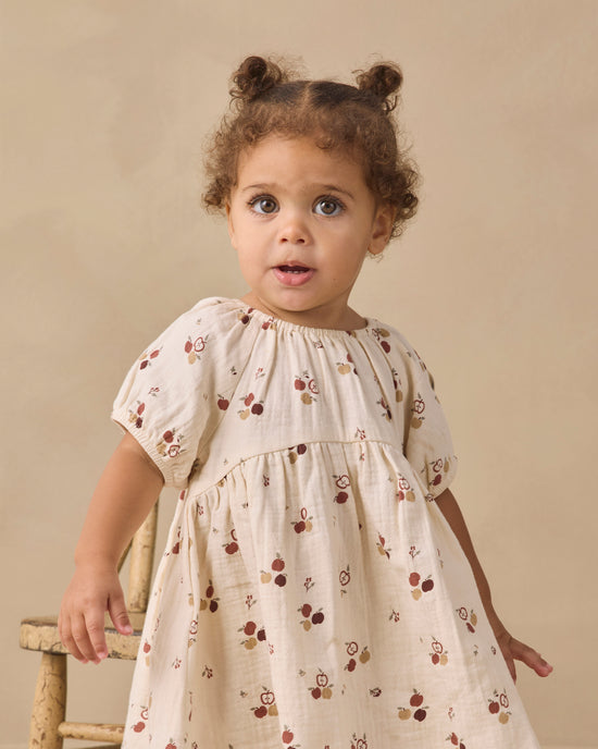 A toddler girl with two buns in her hair wears a cream-colored short-sleeve dress with a brown and red acorn pattern, standing next to a rustic wooden chair against a beige background.