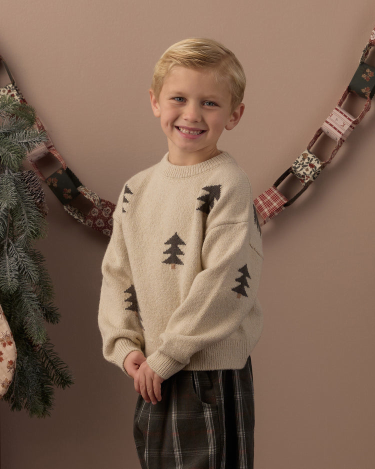 A young blonde boy smiling while wearing a cream knit sweater with black pine tree motifs and dark plaid pants, standing against a warm brown wall with a holiday garland in the background.