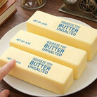 Three blocks of 'Squeeze Toy Butter Unsalted' on a plate with a hand touching one of them.