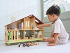 A young child sits on the floor playing with the Wooden Cedar Chalet Modernist Dollhouse by Mentari, placing miniature figures and furniture inside the open-front mid-century modern home while additional wooden dolls and pieces rest on the floor nearby.