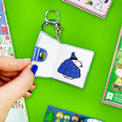 Keychain with a small book design featuring a character in a blue coat and hat on a green background.