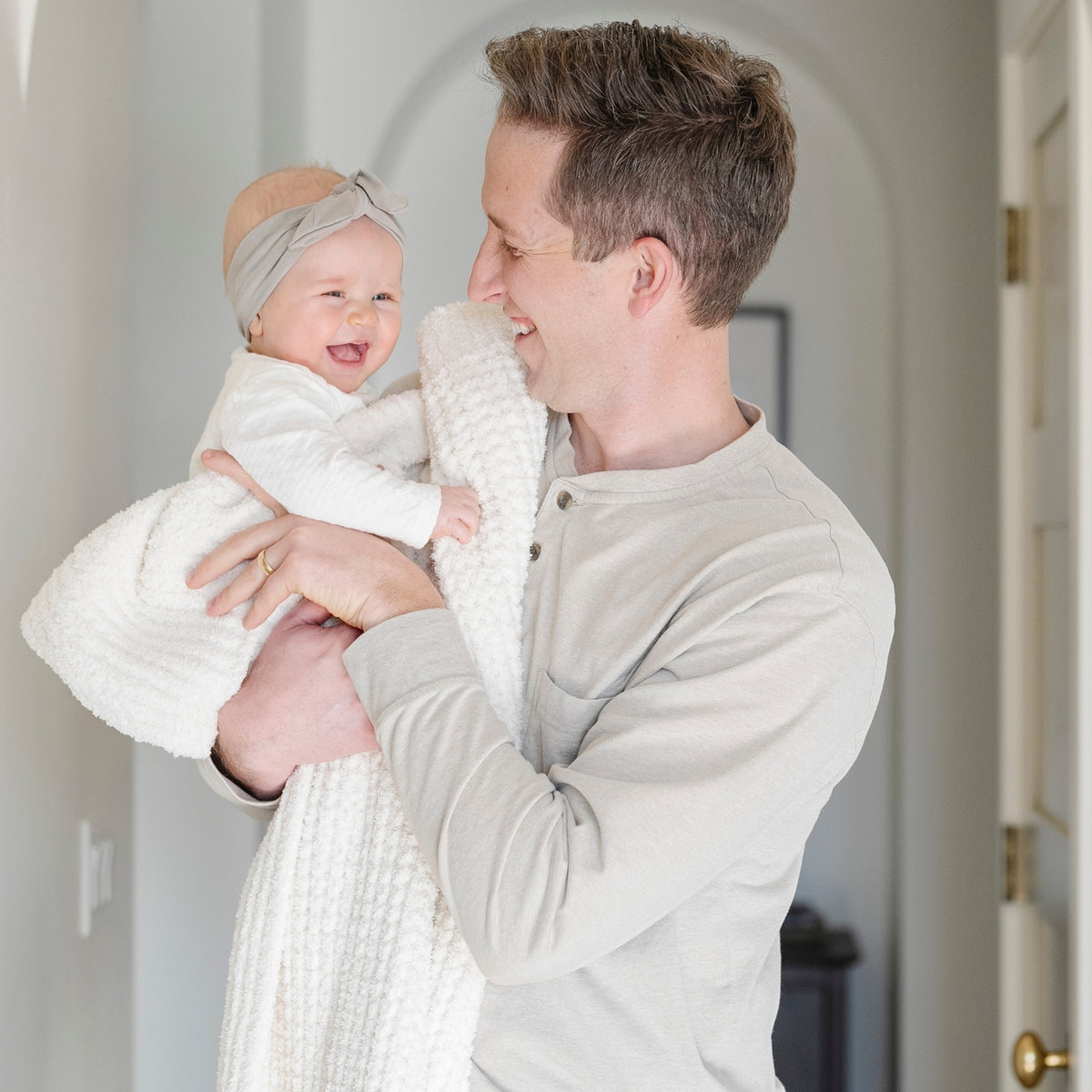 A smiling man in a light gray long-sleeve henley shirt holds a baby wrapped in a white textured blanket. The baby wears a white outfit and a soft beige headband.