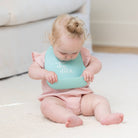Baby wearing a light blue bib with text, sitting on a white surface.