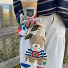 Person holding a drink with a beach background, wearing a striped sweater and white pants.