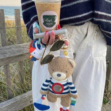 Person holding a drink with a beach background, wearing a striped sweater and white pants.