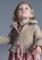 Child wearing a beige ribbed sweater over a plaid dress against a gray background