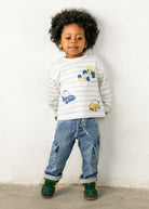 Child wearing a striped shirt with car designs and blue jeans against a white wall.