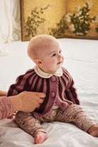 A baby sitting on a white bed wearing a burgundy and brown striped knit cardigan with a cream Peter Pan collar and patterned pants, while an adult hand adjusts the cardigan.