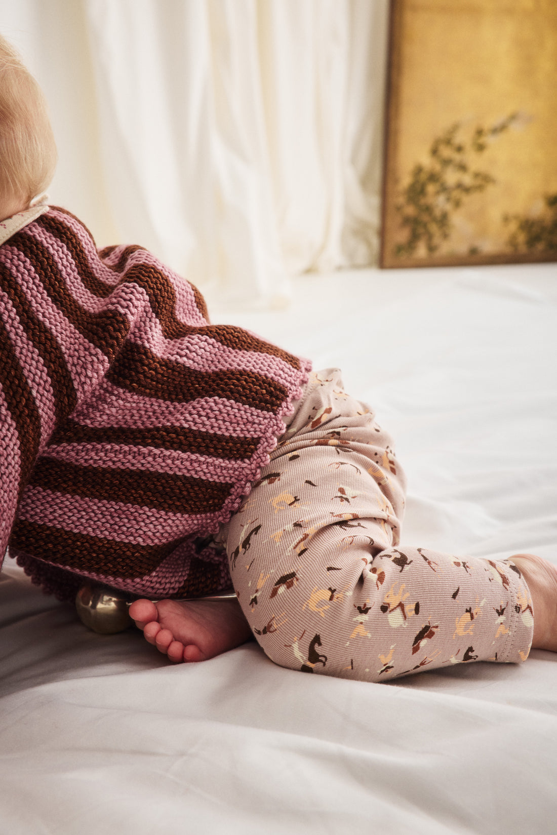 A baby wearing a pink and brown striped knit top and beige patterned leggings, lying on a white bed.