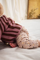 A baby wearing a pink and brown striped knit top and beige patterned leggings, lying on a white bed.